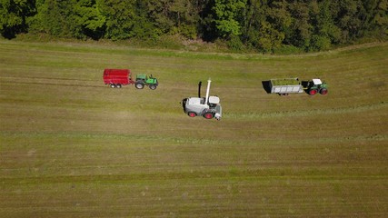 Obraz premium Landwirtschaft: Feldhäcksler, Traktor mit Ladewagen bei Aufnahme von Gras für Grassilage auf einer gemähten Wiese, Vogelperspektive