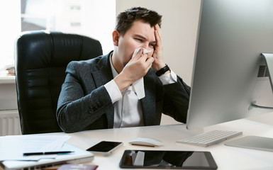 A young businessman or office worker sneezes, coughs, blows his nose at the workplace. Overworked worker got colds and flu. High temperature with ARVI or SARS. Epidemic of influenza or coronavirus.