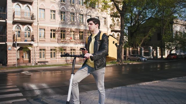 Delivery man with yellow bagpack standing with electric scooter in city and using smartphone to find the client's adress