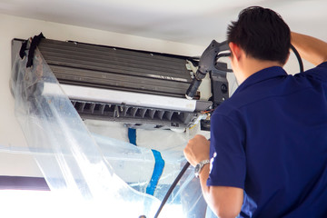 The technicians are cleaning the air conditioner by spraying water. Hand and water spray are cleaning the air conditioner on with white wall background.