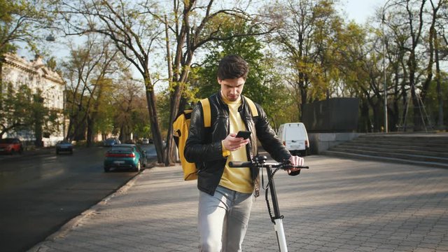 Delivery man with yellow bagpack standing with electric scooter in city and using smartphone to find the client's adress