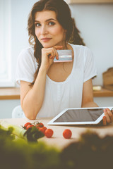 Young woman looking for a new recipe for cooking in a kitchen. Housewife is making online shopping by tablet computer and credit card