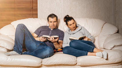 Couple of adults sitting together on sofa and reading books from comfort of their home during covid 19 or coronavirus pandemics and quarantine