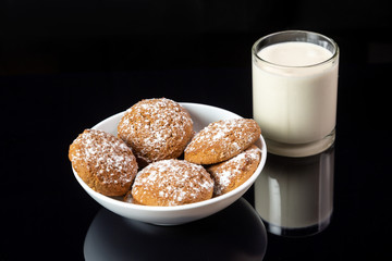 Oatmeal cookies sprinkled with powdered sugar in a white plate and a transparent cup of milk on a black mirror background. Horizontal orientation. 