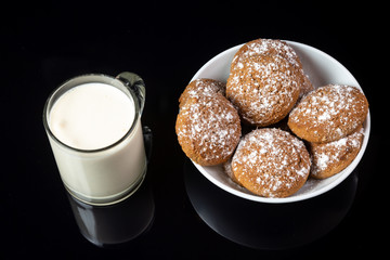 Oatmeal cookies sprinkled with powdered sugar in a white plate and a transparent cup of milk on a black mirror background. Horizontal orientation. 