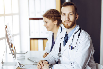 Red-bearded doctor with female colleague discussing current disease therapy while sitting at working place in sunny clinic. Team work in medicine