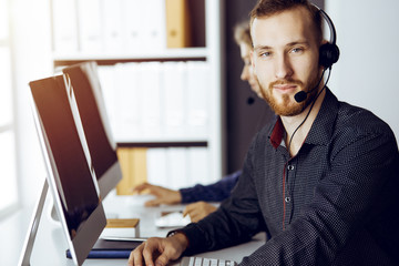 Fototapeta premium Red-bearded businessman talking by headset near his female colleague while sitting in modern sunny office. Diverse people group in call center