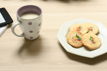 Closeup de uma xícara com leite e biscoitos simples com confeitos coloridos, ao lado um leitor de ebook sobre um caderno.