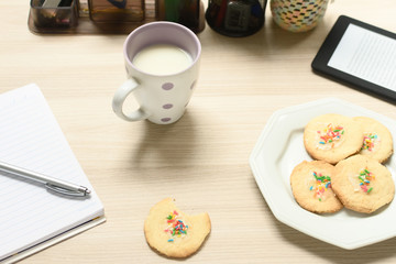Leite e biscoitos simples com confeitos coloridos, um biscoito sendo degustado, cercados por materias de estudo, como caderno, caneta e um leitor de ebook sobre a mesa.