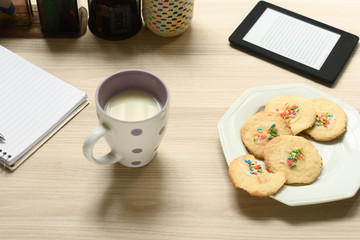 Leite e biscoitos simples com confeitos coloridos, cercados por materias de estudo, como caderno, caneta e um leitor de ebook sobre a mesa.