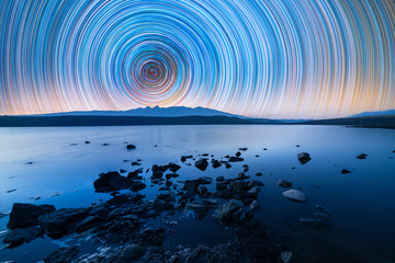 Beautiful night landscape. The shore with stones on the lake and mountains The colorful star trails on the sky. Night time lapse photography.