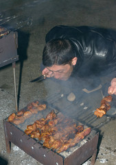Mature man is cooking barbecued meat on compact metal mangal with skewers in evening.