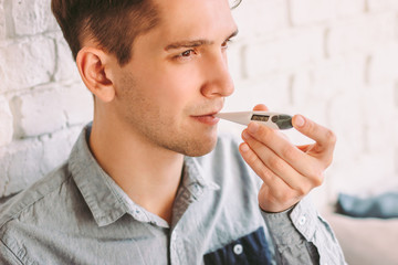 Closeup ill hipster man student putting digital thermometer in mouth for body temperature check....