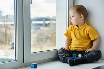 Kid sitting at home on windowsill, playing cubes and watching out the window.