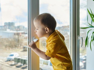 Kid stands at home on window and looks out the window at street through glass.