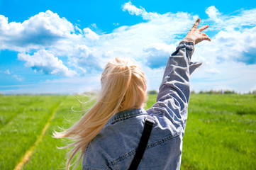 A young girl is looking at the field. Beautiful blue sky. Background green field and sky. The blonde reaches for the sky with her hand in a denim jacket. Beautiful girl admires nature.