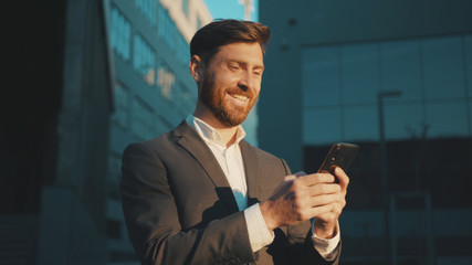 Happy businessman stand in the city center street uses phone texting scrolling tapping smile technology communication sunny day success slow motion