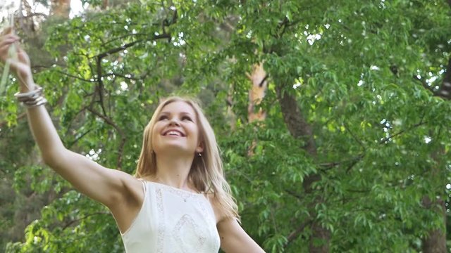 Beautiful attractive girl in a good mood in a white dress in the park admires and waves a dandelion. Slow motion. Green around.
