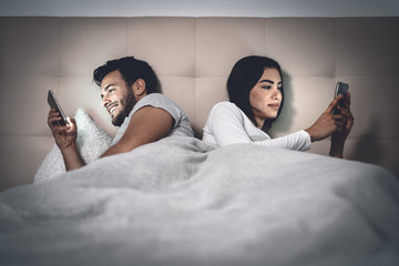 Young couple lying in bed using mobile phone while ignoring each other - Man and woman addicted to...