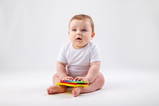 Cute toddler boy 8 months old in white bodysuit plays with developing toy sitting on a white background, space for text