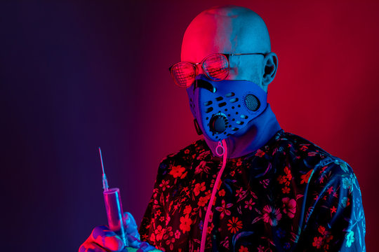 Stylish Bald Man Wearing Respirator Mask And Glasses And Holding Syringe With Vaccine Against Covid 19 In His Hand Over Red Blue Background. Coronavirus Concept.