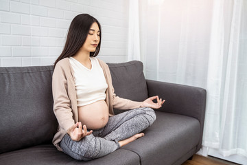 Obraz premium beautiful asian pregnant woman meditating to cope with stress, ache and pain on pregnancy hormones, meditation sitting on sofa hand on knee position, with white brick wall background and a plant pot