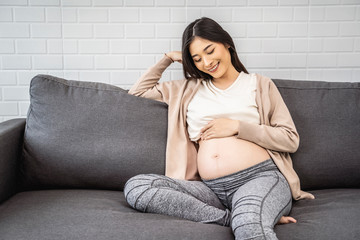beautiful asian pregnant woman placing hands on baby lump feeling heartbeat of baby smiling joyfully, sitting on sofa relaxing resting from tiredness, living room with brick texture wall  