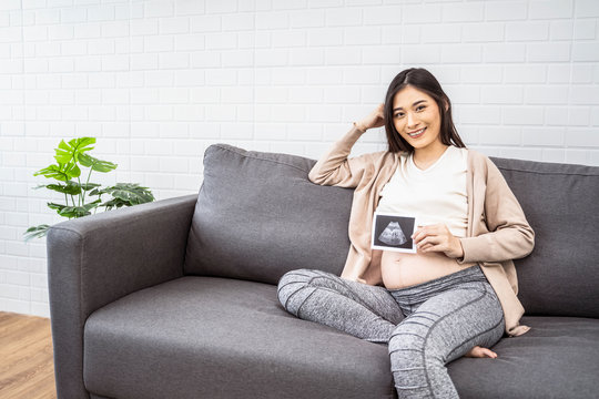 beautiful asian pregnant woman sitting on sofa holding ultrasound baby picture and smiling with happiness, resting relaxing in living room from hormone stress, wearing comfy stretch pants and jumper