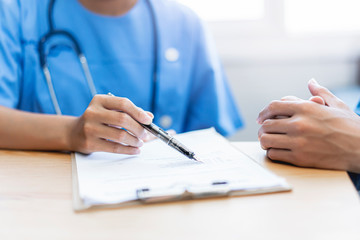 Naklejka premium close up of asian female doctor witting on a documentation on a clipboard on diagnosing patient health or filling in a medical verification form of the patient, sitting in hospital office medical room