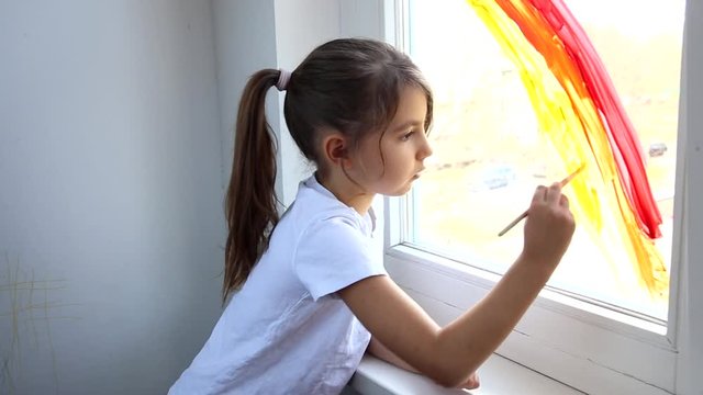 Little girl painting rainbow on window during Covid-19 quarantine at home. Chase the rainbow flashmob and staying at home concept