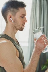 Young man in earphones and sports vest holding bottle of water by his mouth