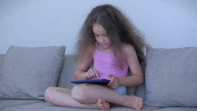 Curious cute girl preschool child using a digital tablet technology device looks telephone.Plays in the phone sitting on the sofa alone.Children tech addiction concept play game at home.