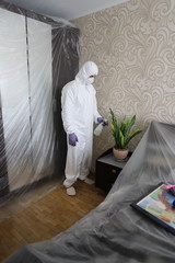 a man in a protective suit in an apartment sprays a live green home flower in a pot, life in an epidemic,