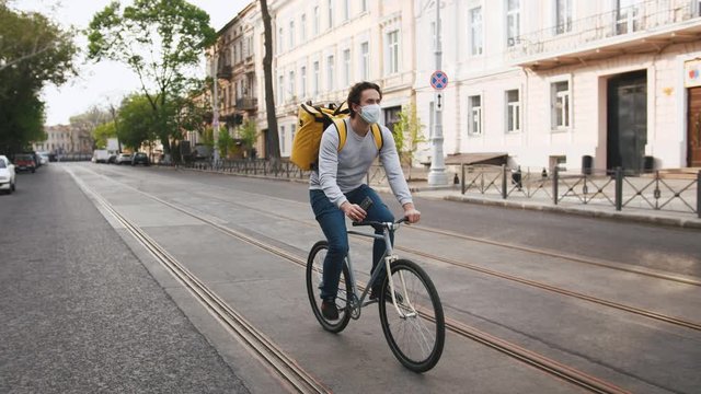 Courier with yellow backpack and protective mask rides a bicycle on the street with food delivery and using smartphone in the morning