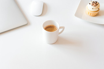 Cup of coffee and cupcake, silver laptop on white desk. Coffee break in home office concept. 