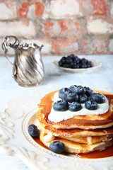 A stack of blueberry pancakes on a white textured plate. In the distance, a vintage gravy boat. Copy of the space.