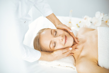 Fototapeta na wymiar Beautiful happy woman enjoying facial massage with closed eyes in spa salon. Relaxing treatment in medicine and Beauty concept