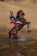 A young adult man in knightly armor rides a horse on a river along a sandy shore
