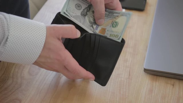 Male hands puts money hundreds dollar bills into leather wallet at work table, close-up view
