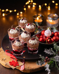 Dessert with red fruits sauce, chocolate mouse, sour cream in glasses. Wooden background with garland lights bokeh and christmas decoration. Christmas, new year holidays background concept.