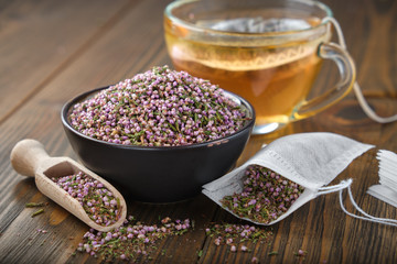 Bowl of dry healthy heather, tea bag with Erica flowers. Glass tea cup with herbal teabag inside on wooden table. Herbal medicine.