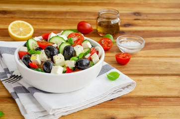 Greek salad of fresh juicy vegetables, feta cheese, herbs and olives in a white bowl on a wooden background. Healthy food. Horizontal orientation, copy space