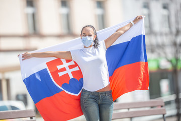 Smiling girl in a face mask holds a flag of Slovakia behind her on the street