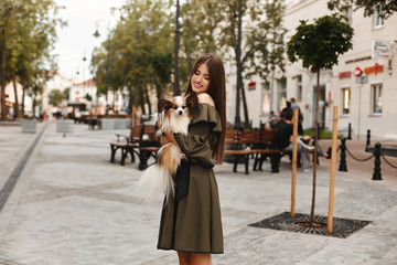 Beautiful model girl with broad smile in short dress posing with small cute papillon dog on her hands outdoors at the old city center. Summer fashion