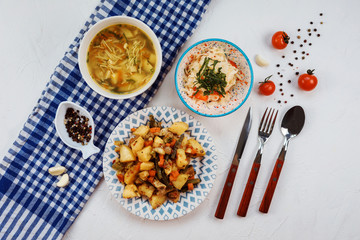 Delicious lunch set on the white table with blue towel on it. Lunch set contains coleslaw, potatoes with meat and noodle and potato soup. Table is decorated with pepper, garlic and tomatoes. 