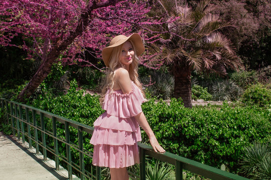 Beautiful blonde girl in a hat and a pink dress walks in a flowering garden.