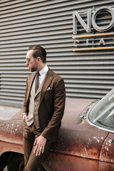 Outdoor fashion photography of a man wearing brown business suit and tie leaning on the vintage car. Concept for man’s magazine cover or content.