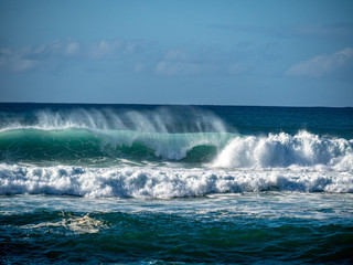 Big waves crash close to shore with aquamarine seas, white foam, sand and blue skies.
