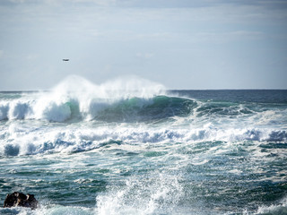 Naklejka premium Big waves crash close to shore with aquamarine seas, white foam, sand and blue skies.