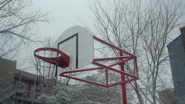 Pan shot of snow covered basketball hoop and net during winter season with heavy snowfall during foggy day with dried tress and building around